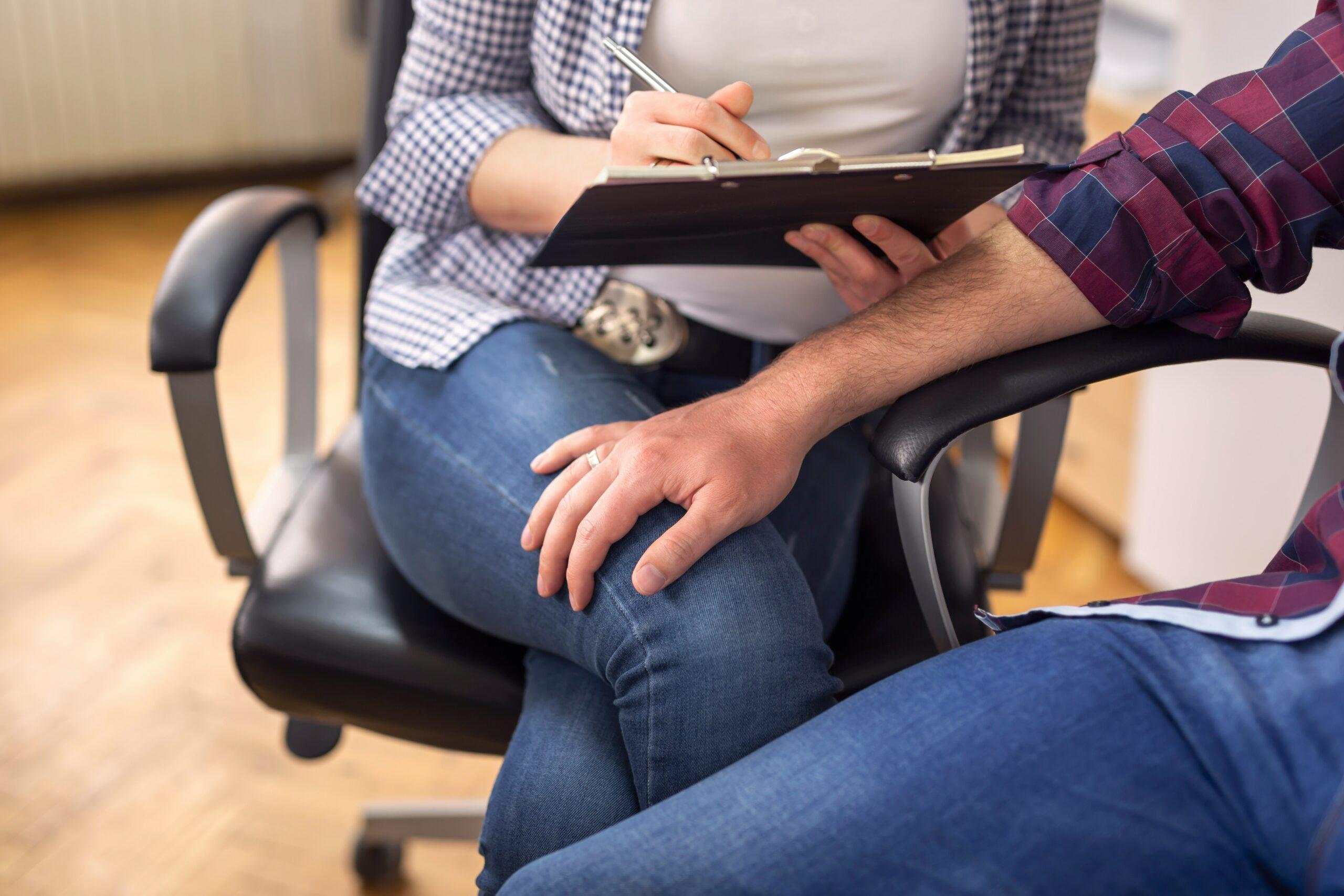 Close-up of a man placing his hand on a woman's knee while she writes notes on a clipboard during a conversation in an office setting.
