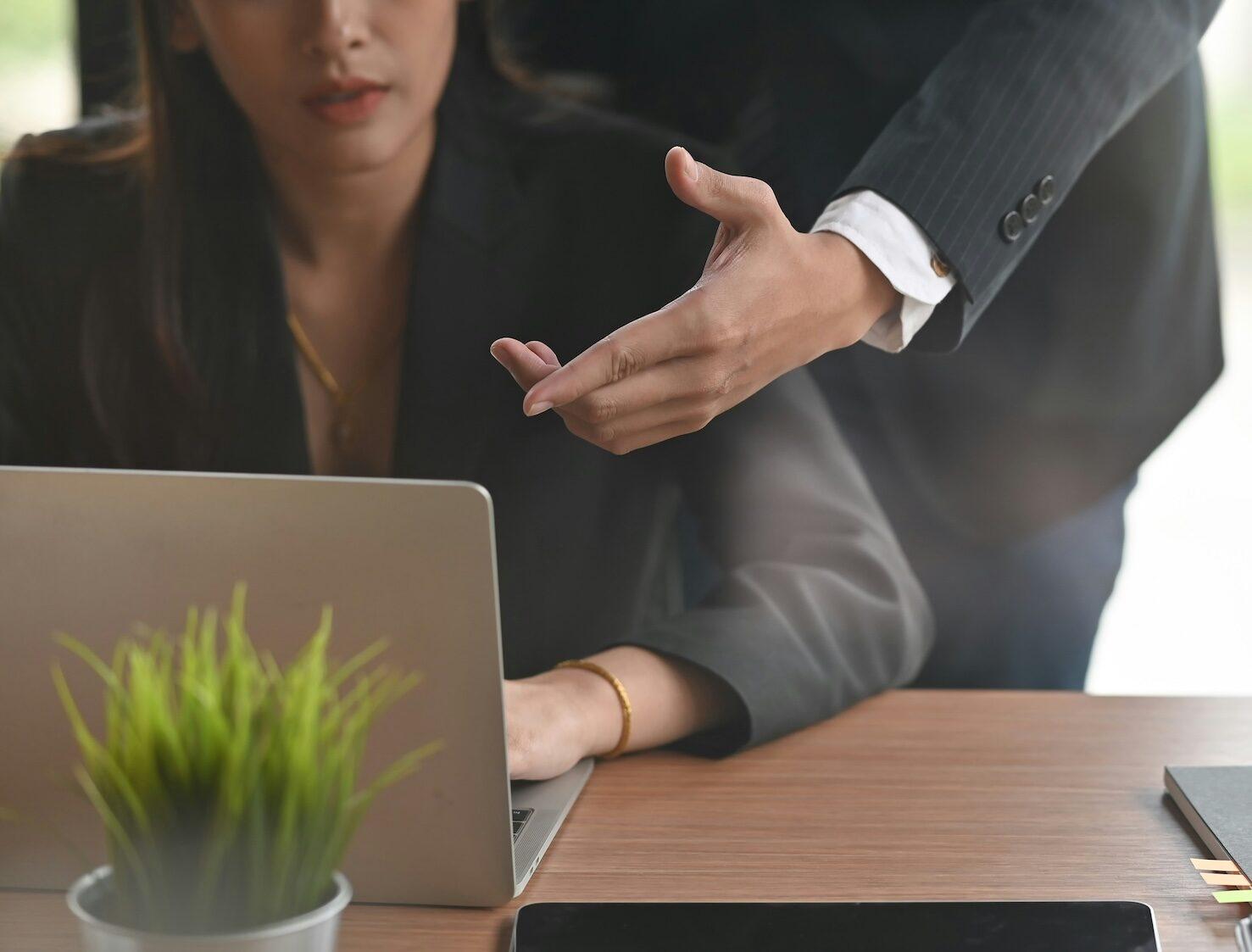 A woman in business attire seated at a laptop computer. A man is standing behind her, reaching over her shoulder and pointing at the screen.