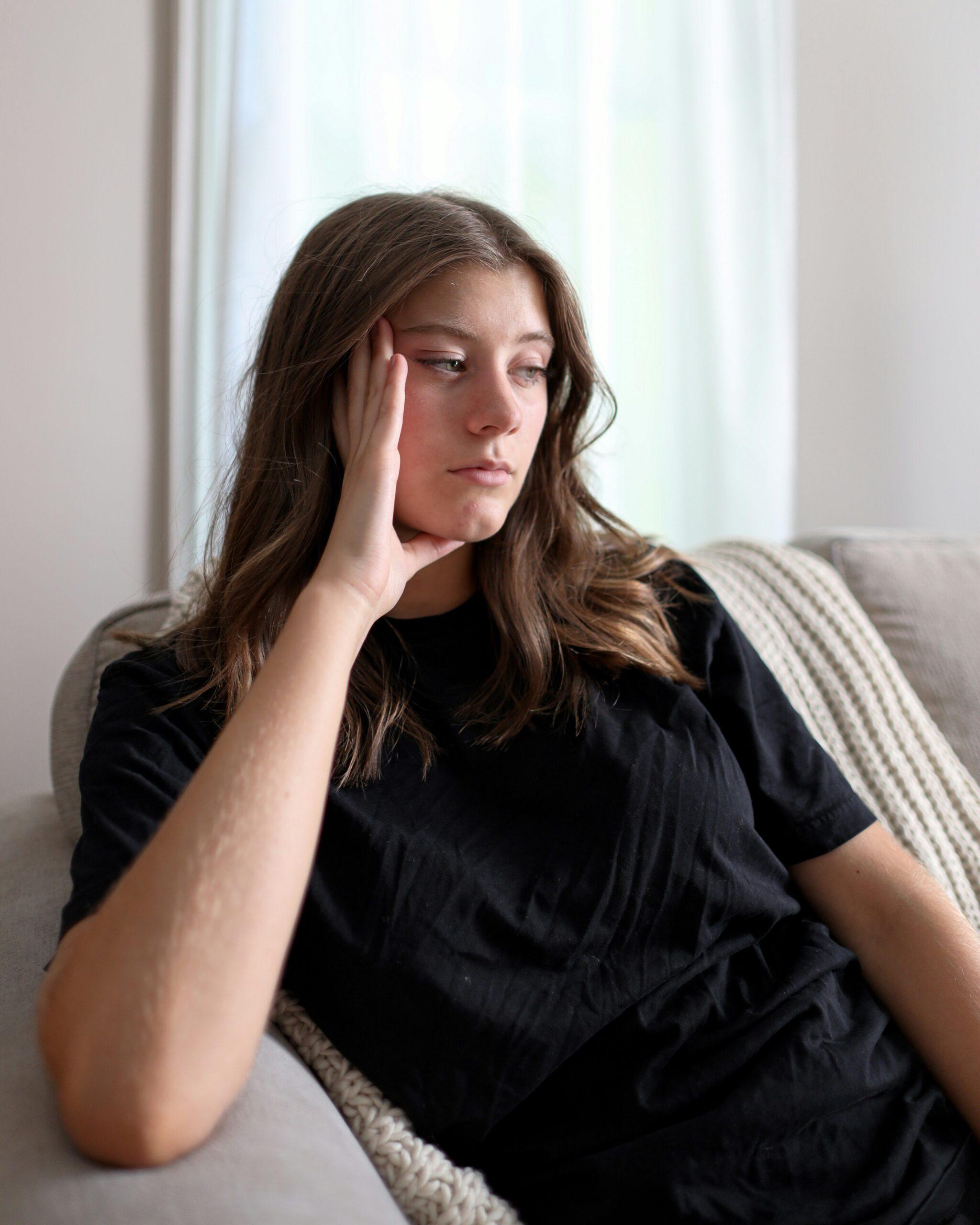 A woman sitting on a couch, resting her hand on her head, appears deep in thought or contemplation.