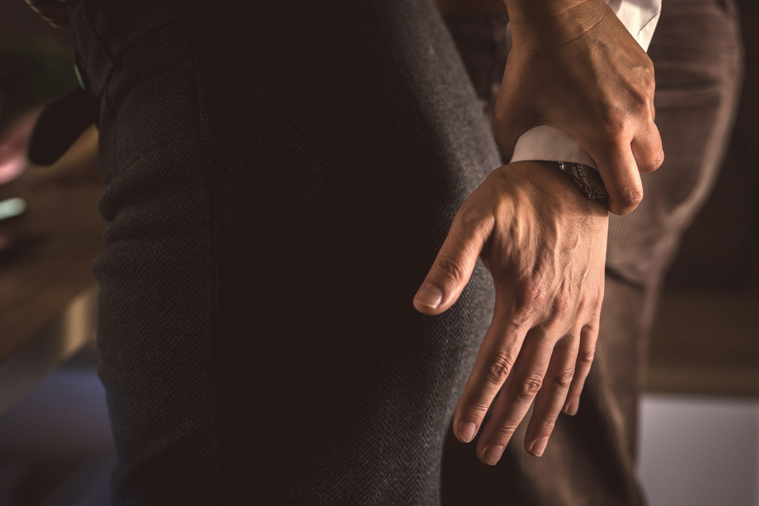 close up of an older man's hand hovering over the behind of a woman in a pencil skirt. She has grabbed his hand to stop him from touching her.
